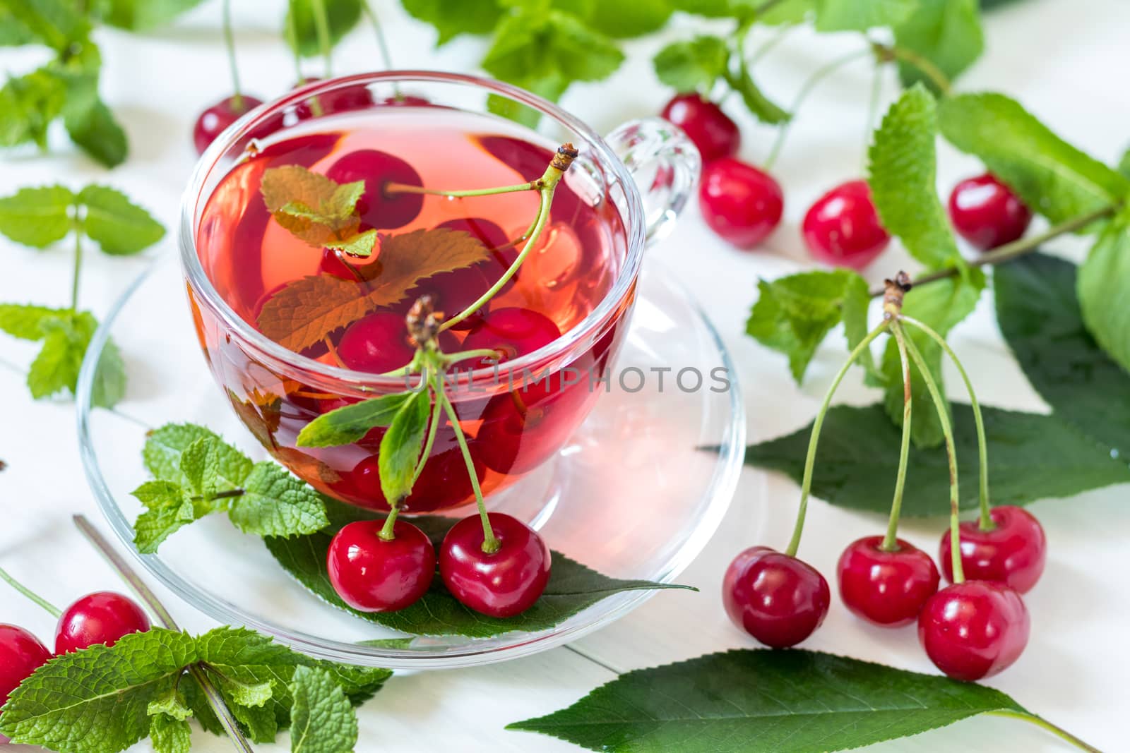 Fresh fruit cherry drink in transparent glass cup surrounded by cherries on the white wooden background.
