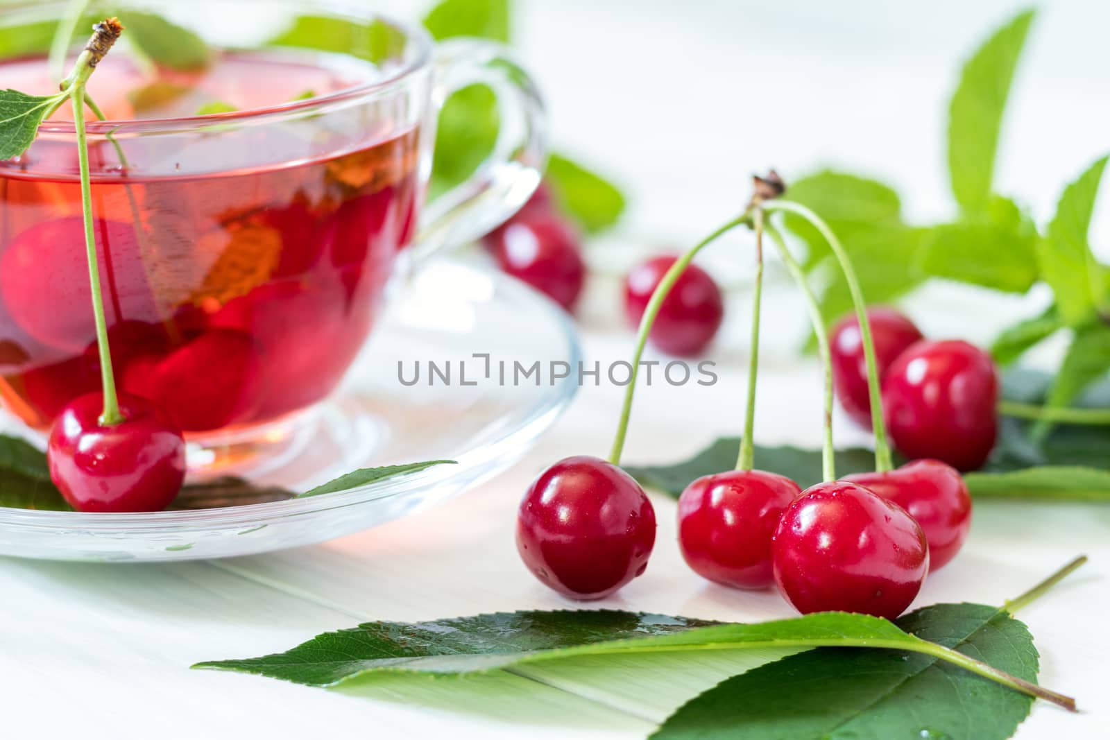 Fresh juicy cherries and cherry flavored drink in transparent glass cup on the white wooden background