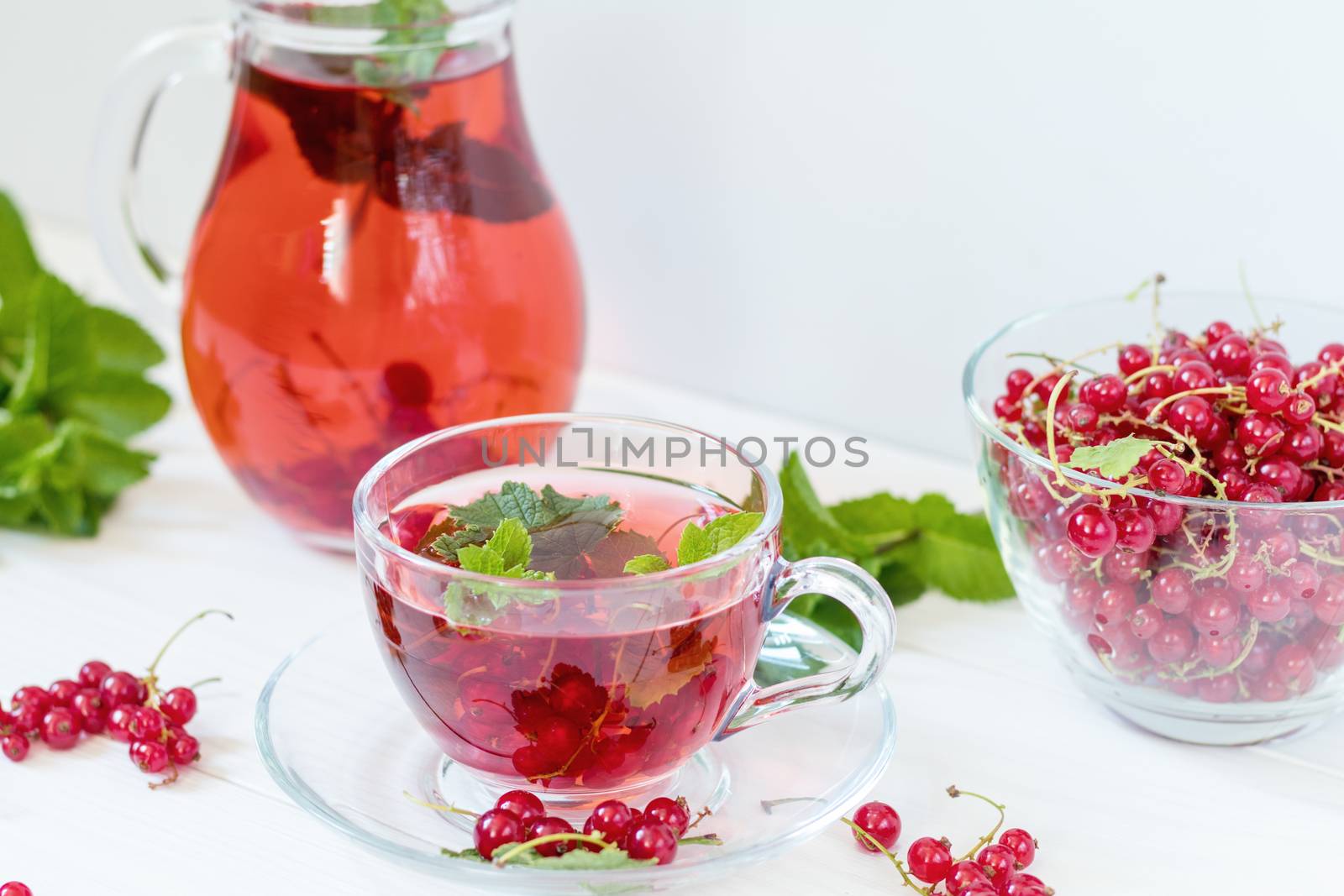 Redcurrant drink in transparent glass carafe and cup. Clear glass vase with red currant berries on the white wooden background
