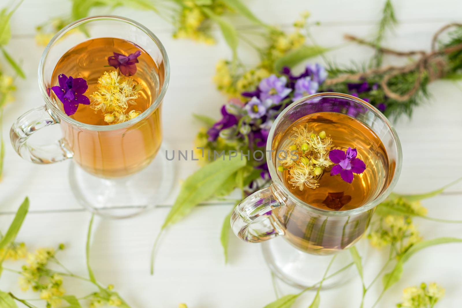 Linden herbal tea in a transparent grog glass with a linden blossom and bunch of herbs on the white wooden surface
