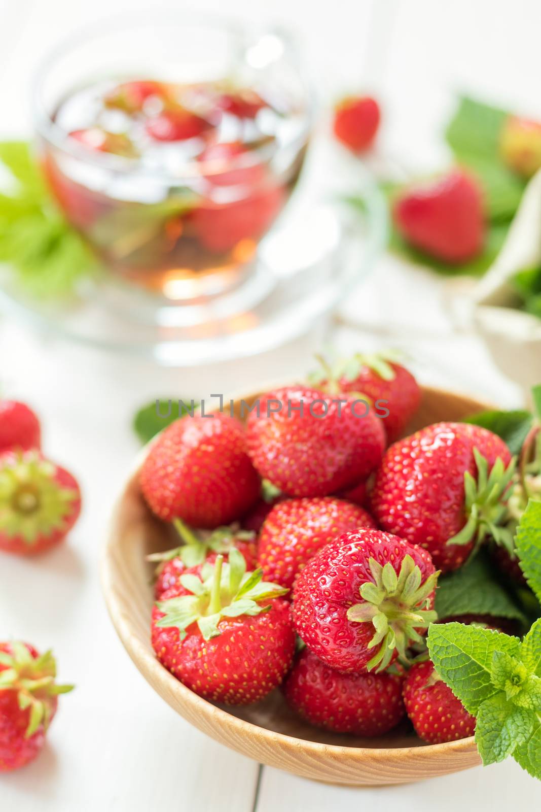 Fresh strawberry in wooden plate. Glass cup of summer tea with fresh strawberry. Green leaves. Fresh mint. White wooden table. Shallow depth of field.