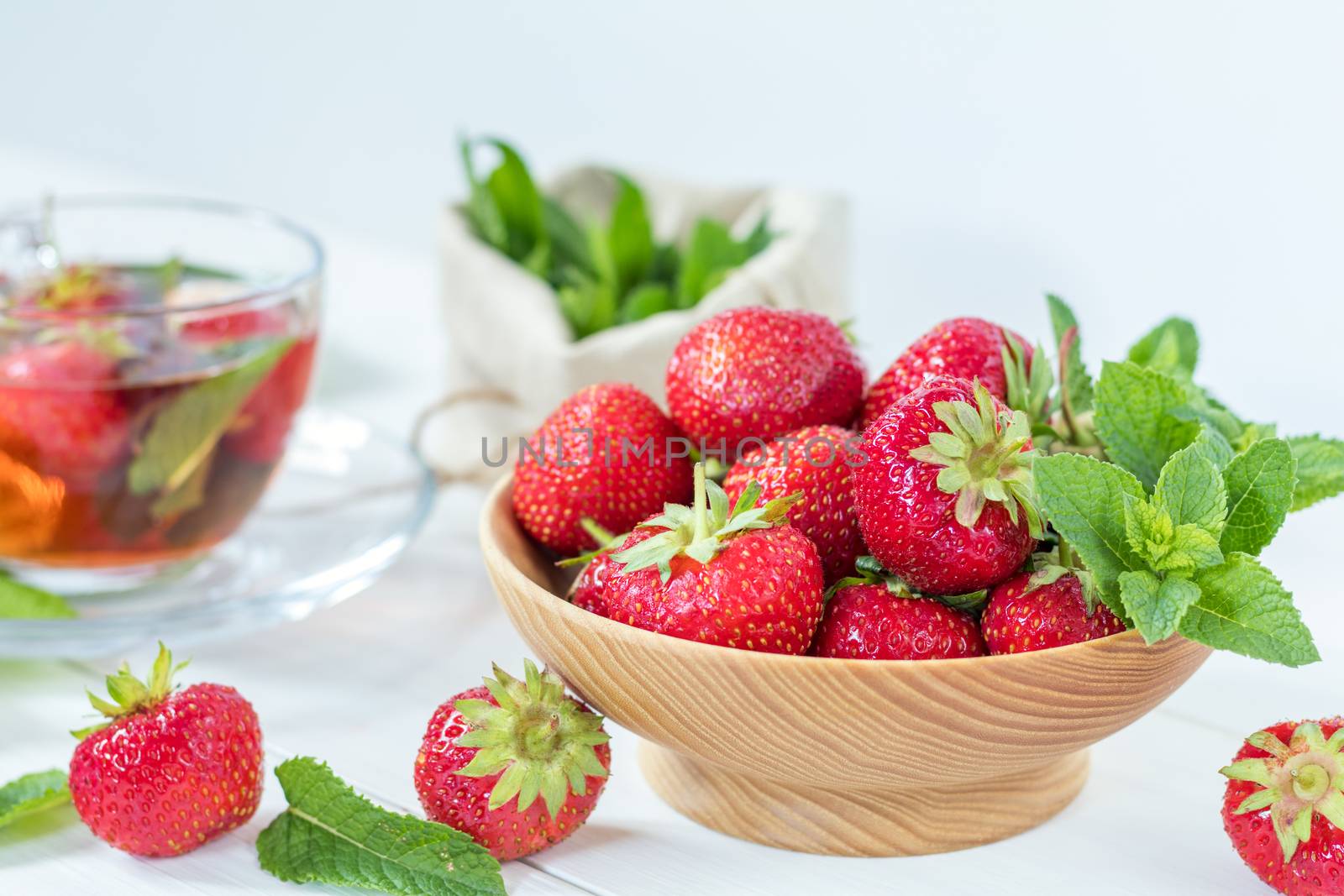 Fresh strawberry in wooden plate. Glass cup of summer tea with fresh strawberry. Green leaves. Fresh mint. White wooden table. Shallow depth of field.