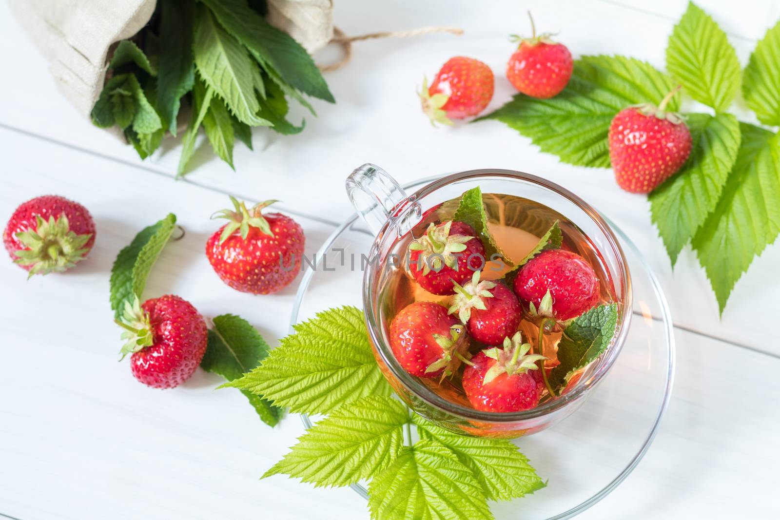 Glass cup of summer tea with fresh strawberry. Green leaves. Fresh mint. White wooden table. Shallow depth of field.