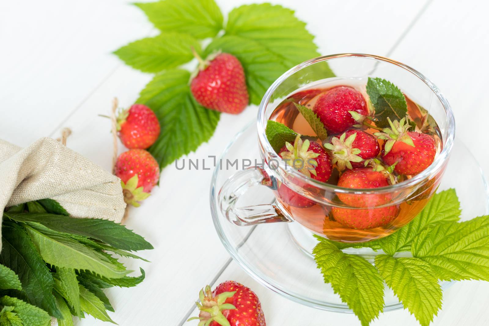 Glass cup of summer tea with fresh strawberry. Green leaves. Fresh mint. White wooden table. Shallow depth of field.