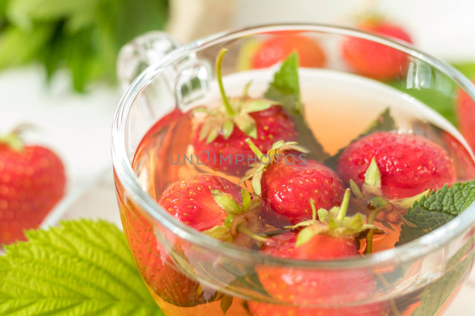 Glass cup of summer tea with fresh strawberry. Green leaves. Fresh mint. White wooden table. Shallow depth of field.