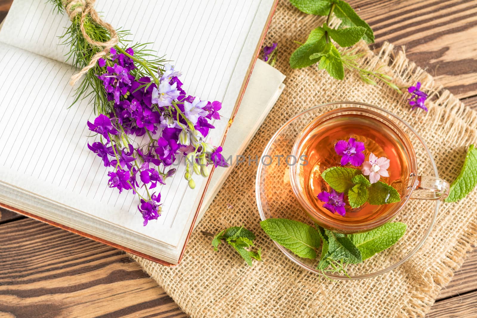 Glass cup of summer herbal tea with fresh mint and field larkspur. Bouquet of wild flowers on the notebook. Wooden table. Shallow depth of field.