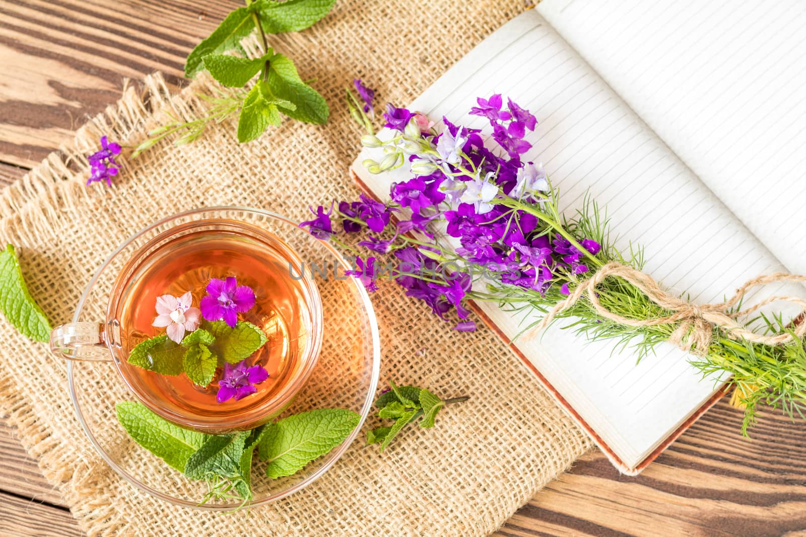 Glass cup of summer herbal tea with fresh mint and field larkspur. Bouquet of wild flowers on the notebook. Wooden table. Shallow depth of field.