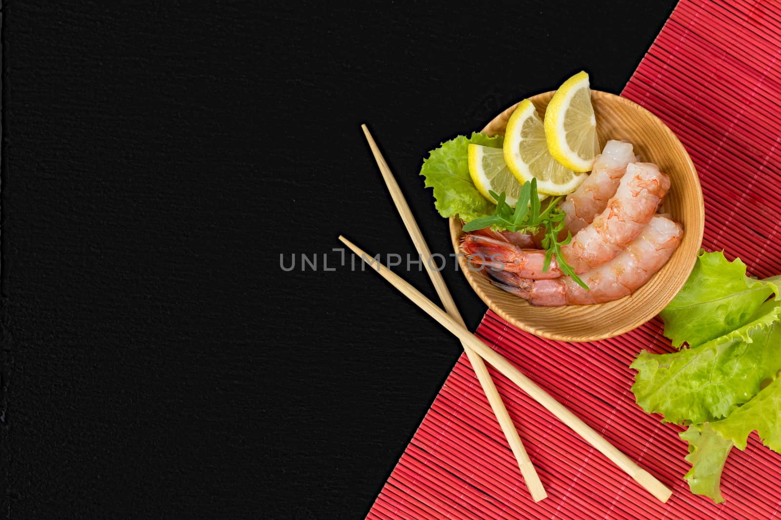 Cooked peeled shrimps with lemon slices, green lettuce and arugula leaves on a wooden plate with wooden chopsticks crossed. The plate, chopsticks and green lettuce leaves are on red makisu