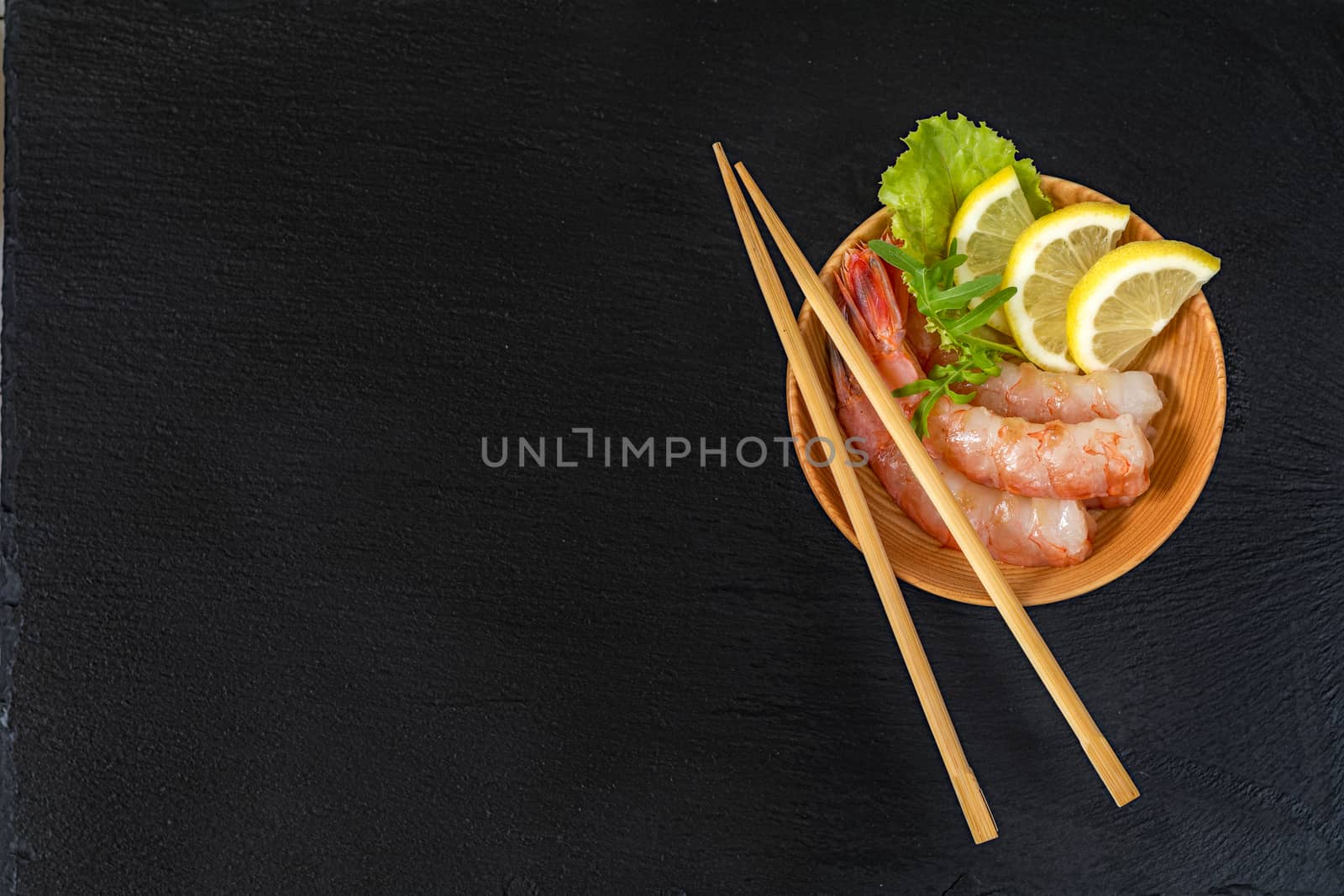 Cooked peeled shrimps with lemon slices, green lettuce and arugula leaves on a wooden plate with wooden chopsticks on a side of a plate. The plate is at the black stone countertop