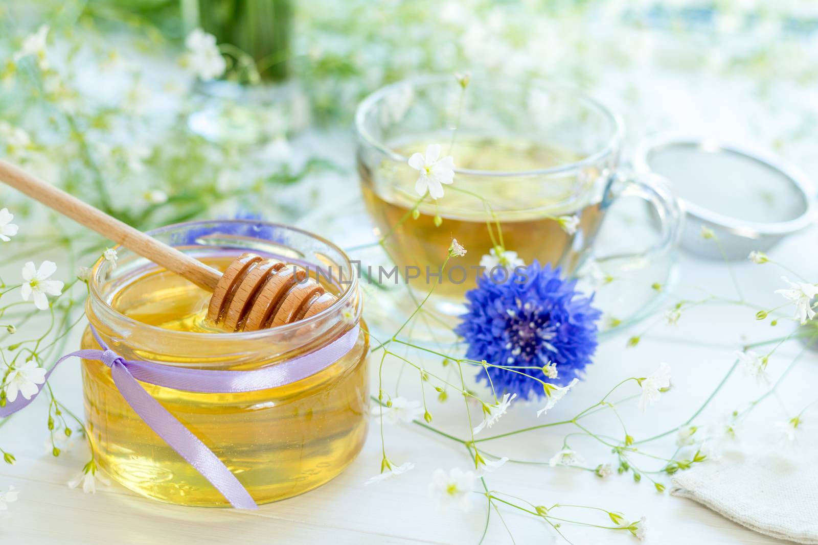 Honey in glass jars and cup of tea with white spring flowers and cornflower on windowsill. Shallow depth of field.