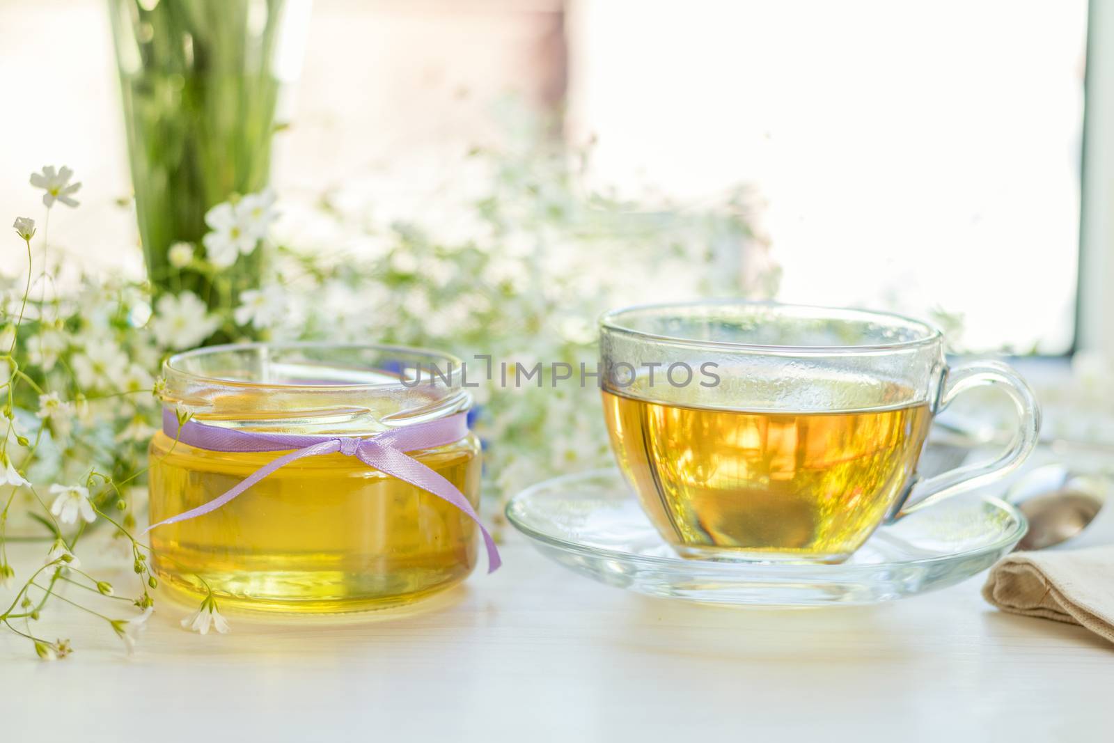 Honey in glass jars and cup of tea with white spring flowers on windowsill. Shallow depth of field.