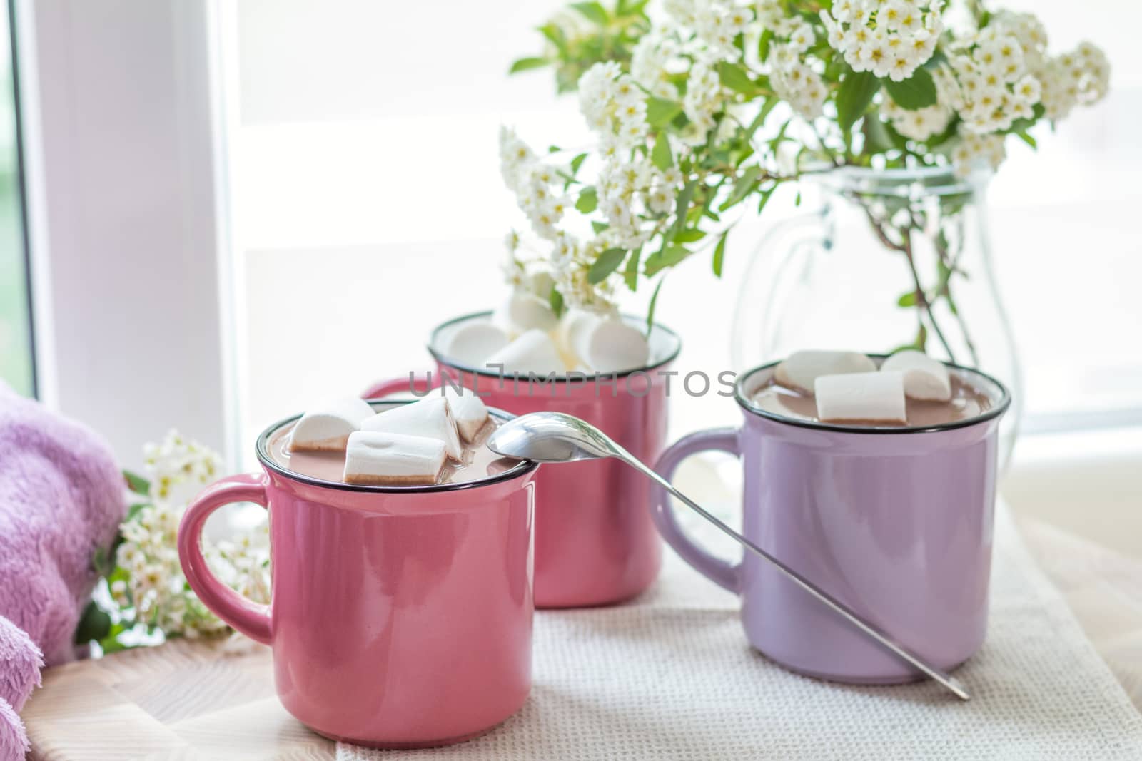 Marshmallows on top of hot cocoa in pink cups on the white napkin on the windowsill. Fresh white spring flowers are framing the image. Cozy home concept. Shallow depth of field