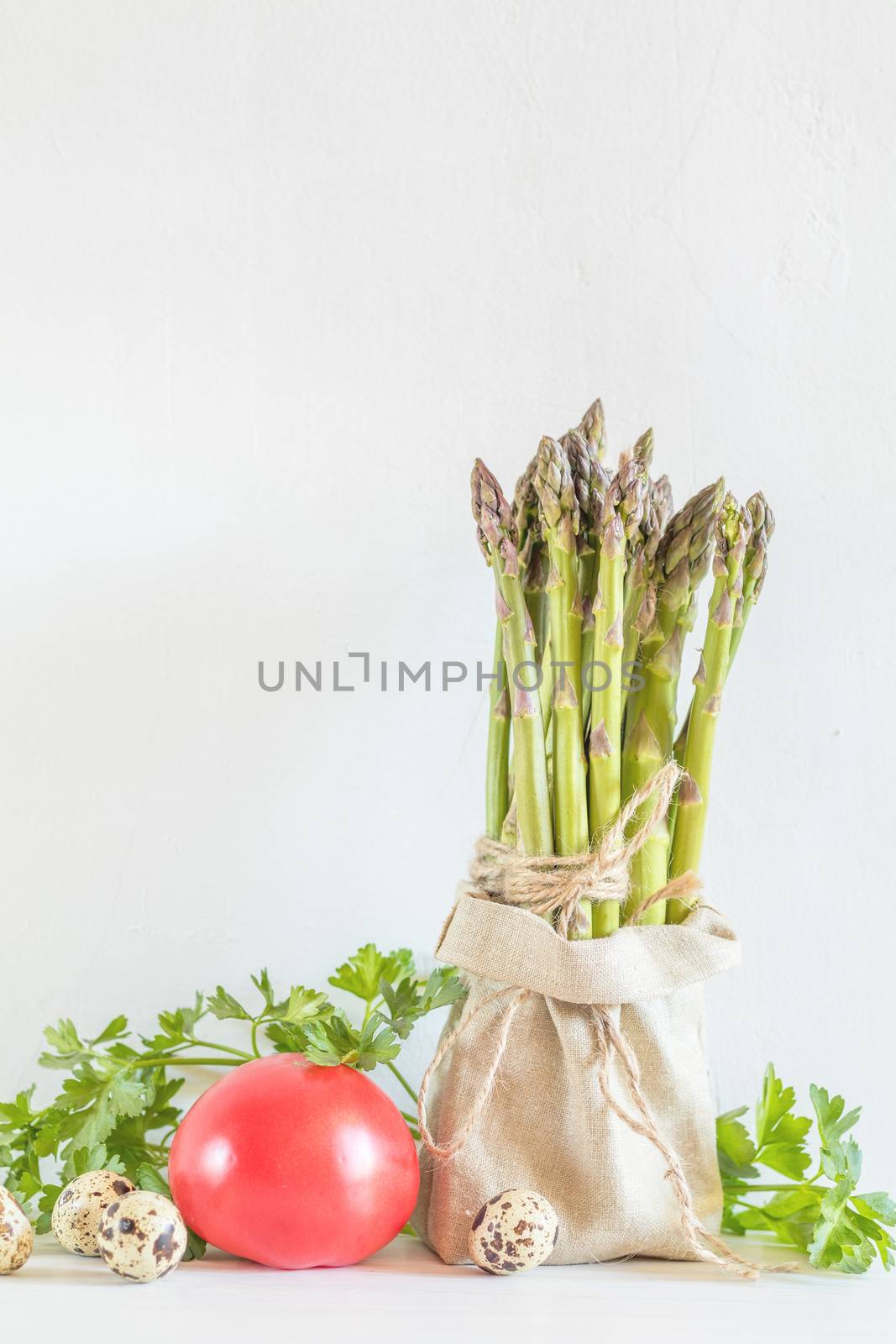 Bunches of fresh asparagus in a little sack, vegetables and quail eggs on the white cracked wall background. Copy space