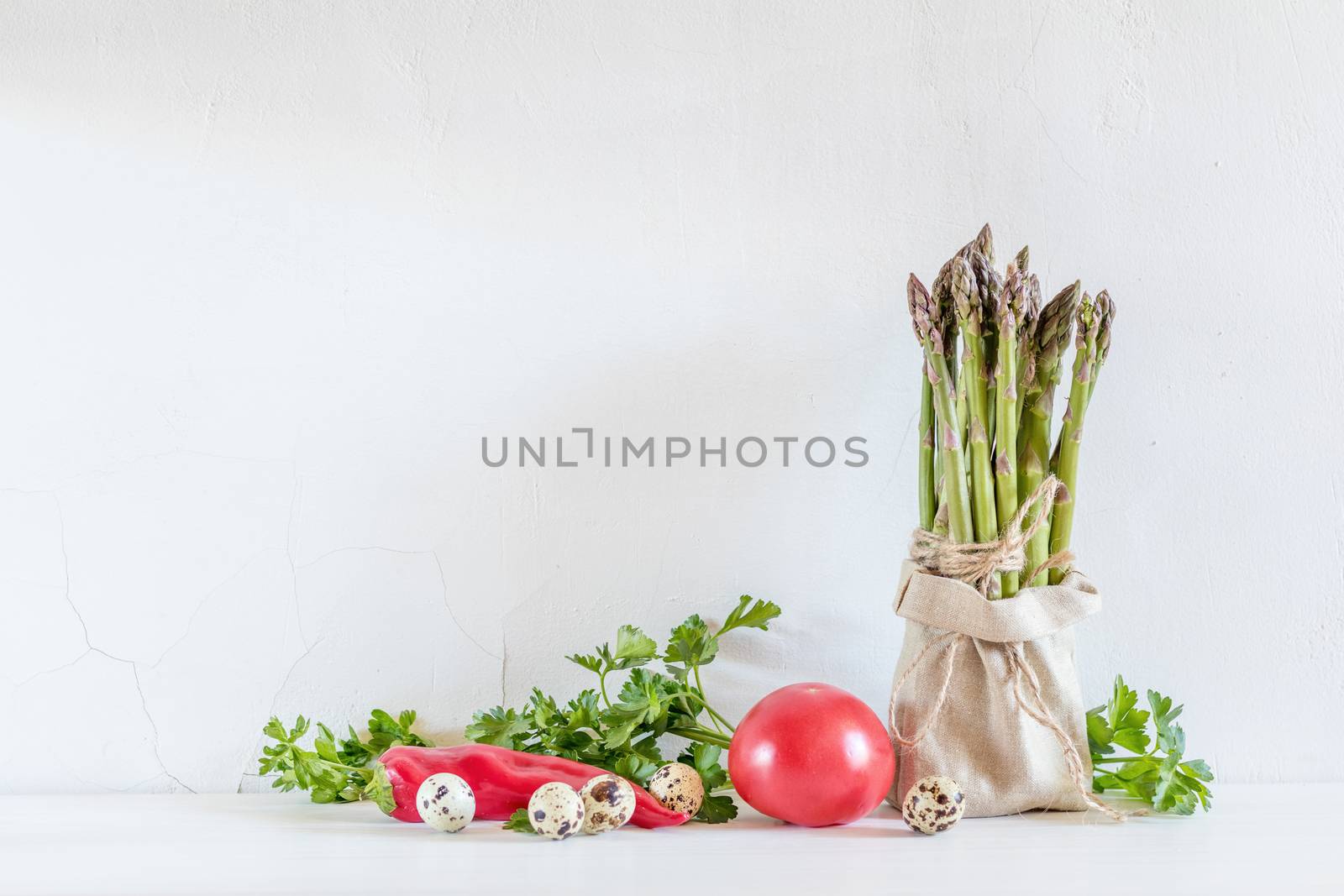 Bunches of fresh asparagus in a little sack, vegetables and quail eggs on the white cracked wall background. Copy space