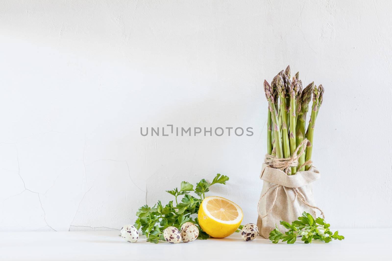 Bunches of fresh asparagus in a little sack, lemon and quail eggs on the white cracked wall background. Copy space
