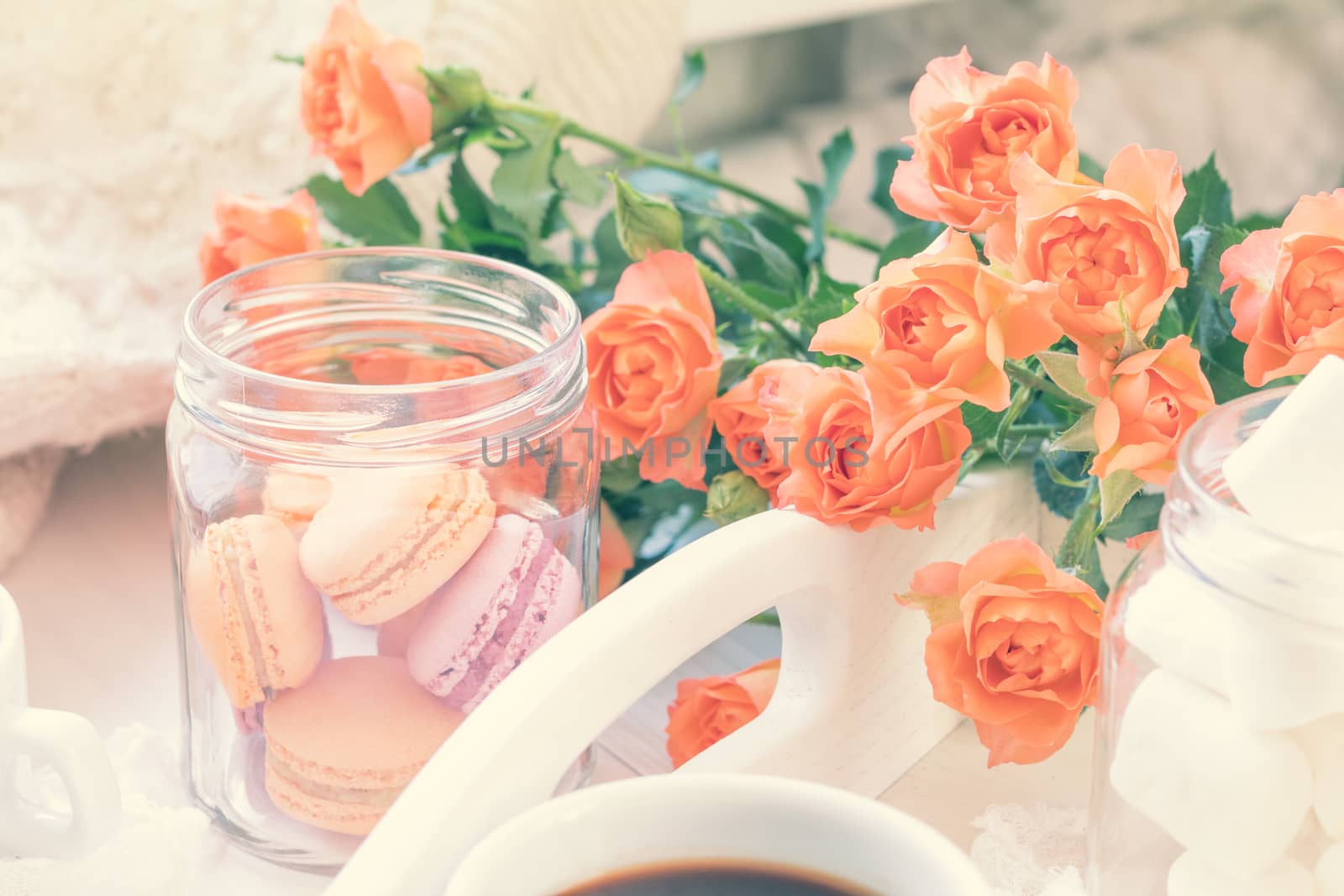 Cup of coffee. Orange mango or citrous macaroons and marshmallow in jars. Fresh little roses. Light wooden background. Sunlight. Toned photo with light vintage style. Shallow depth of field.