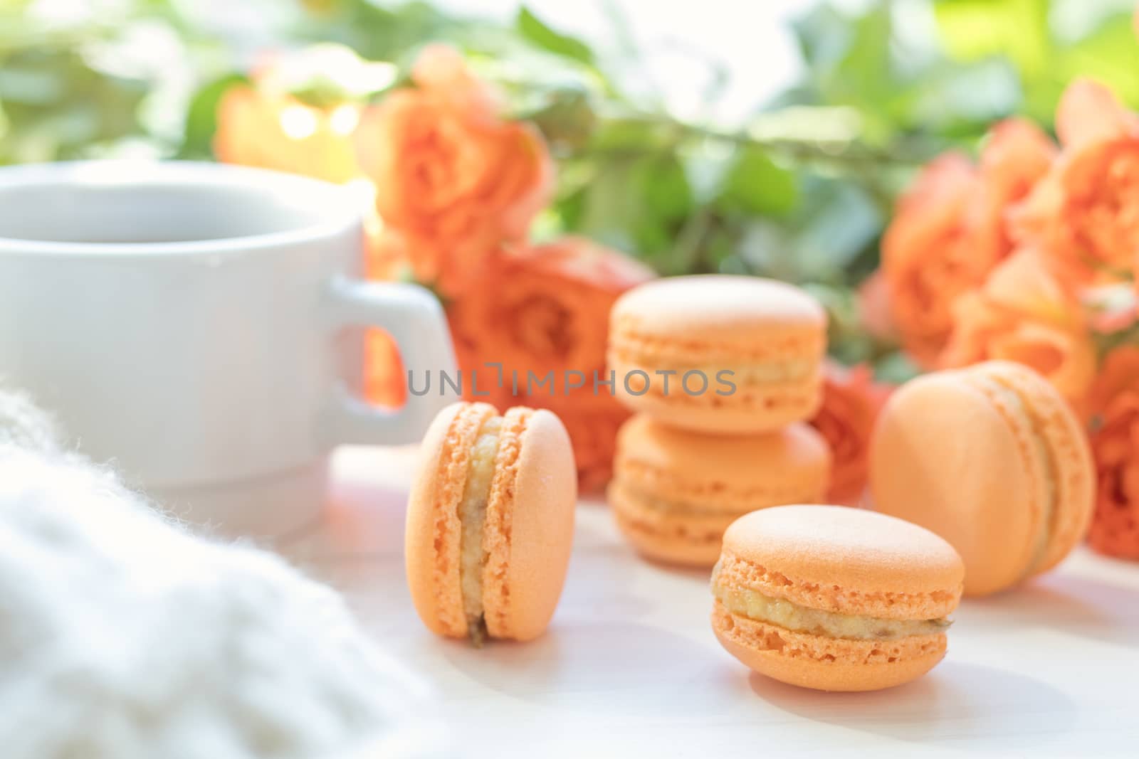 Orange mango or citrous macaroons, cup of coffee and fresh little roses on light wooden background. Sunlight. Coloring and processing photo with light vintage style. Shallow depth of field.
