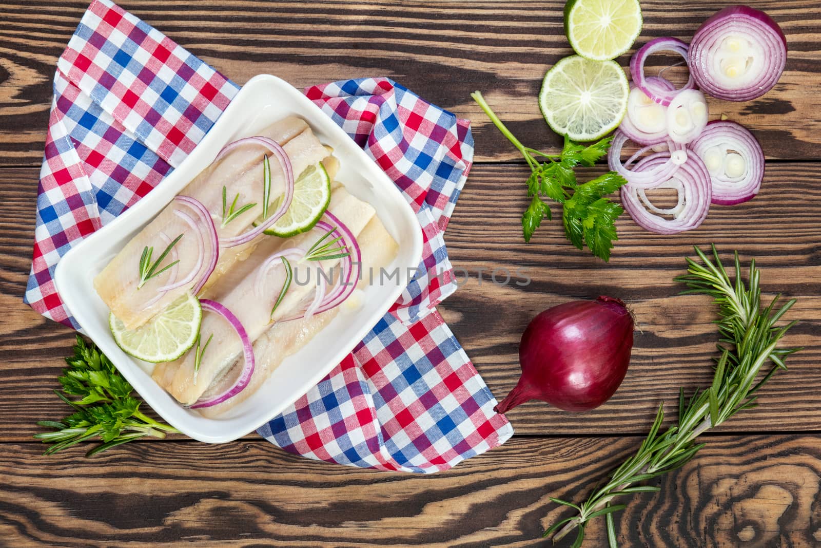 Sliced herring fillets, cut onion and lime on white plate. Checkered napkin. On wooden table.
