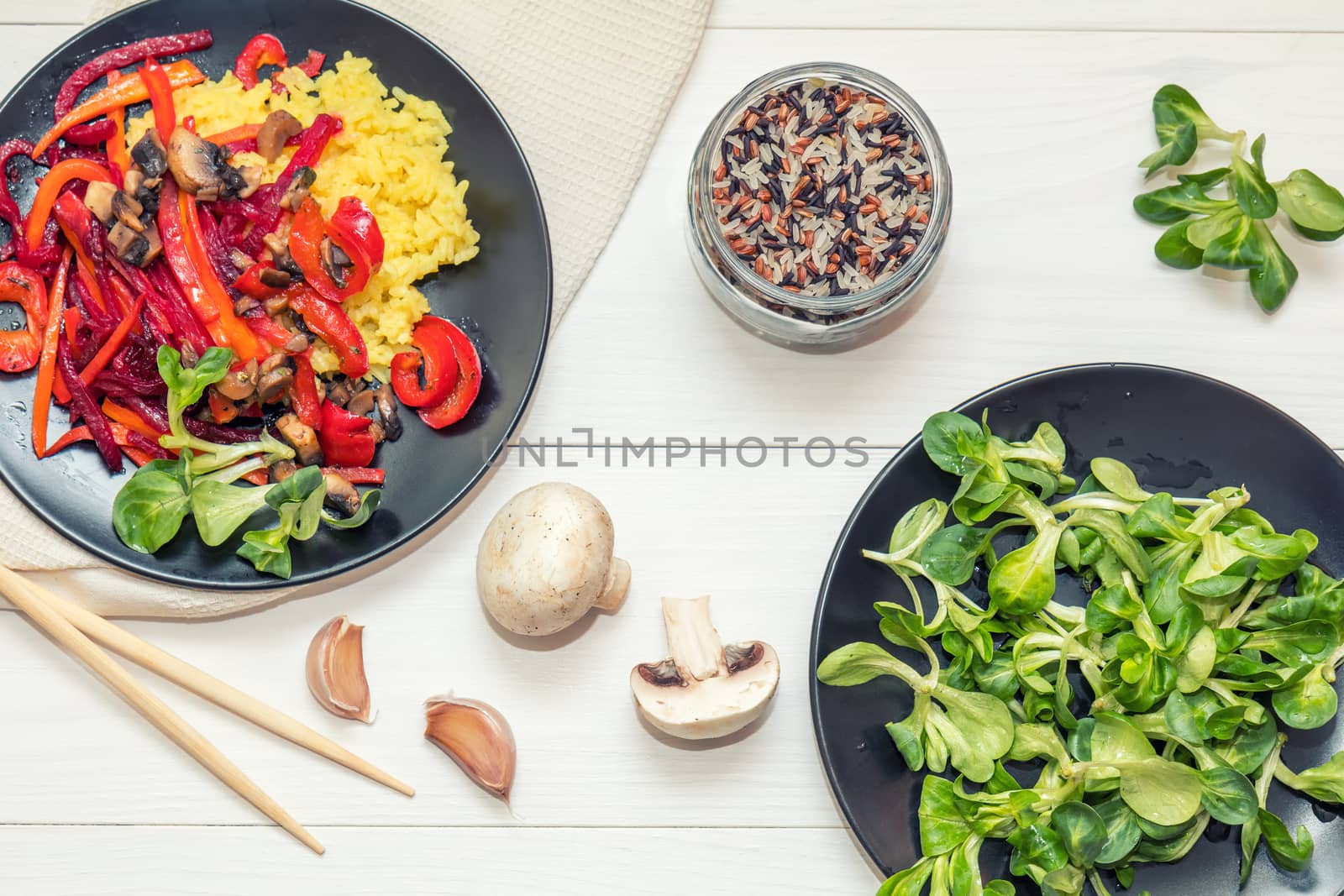 Healthy vegetarian diet concept. Rice and steamed vegetables, lamb's lettuce feldsalat on a black plates, chopsticks, napkin, garlic, mushrooms, raw multicolor rice in glass jar. White wooden table.