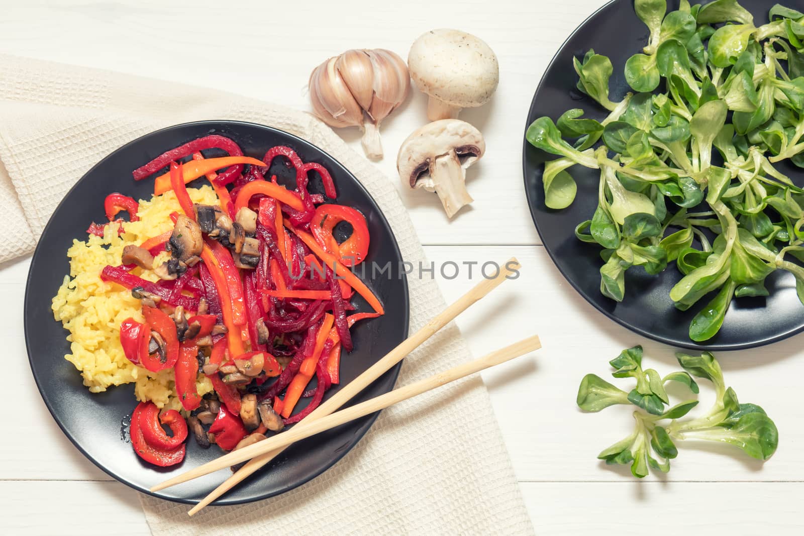 Healthy vegetarian diet concept. Rice and steamed vegetables, lamb's lettuce feldsalat on a black plates, chopsticks, napkin, garlic, mushrooms. White wooden table.