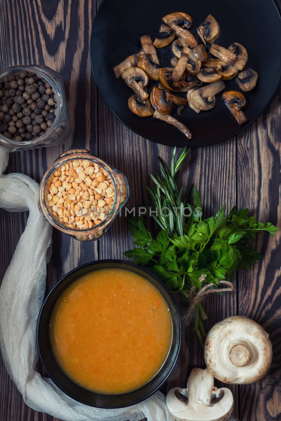 Pea soup in black plate. Fried mushrooms. Dried pea, pepper in glass jars.  Fresh parsley and rosemary. Dark wooden table. Toned.