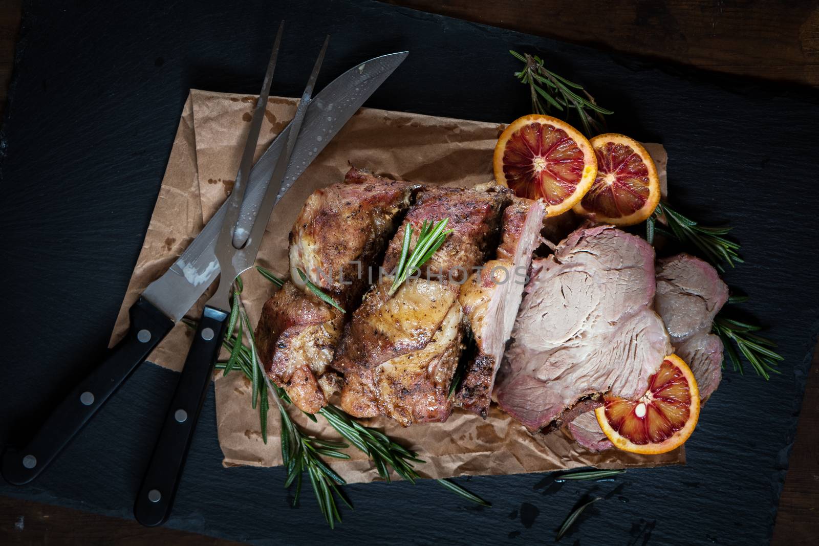 Sliced baked pork with herbs on a stone cutting board on wooden background.Grilled steak on a paper. Rosemary and sliced sicilian blood oranges  on a dark groundwork.