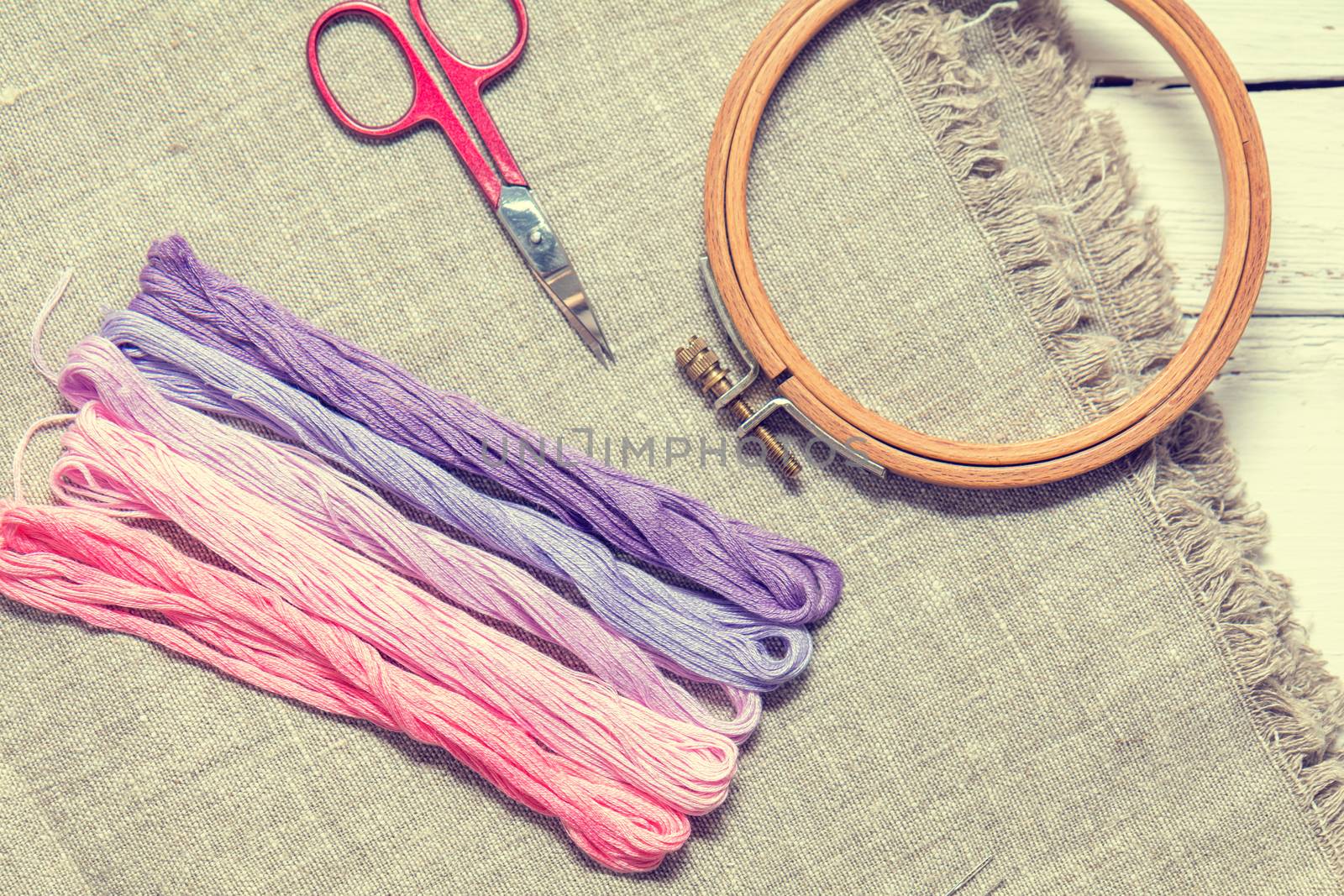 Set for embroidery. Embroidery thread shades of violet and pink color, embroidery hoop, scissors and needle on linen homespun cloth. Coloring and processing photos. Shallow depth of field.