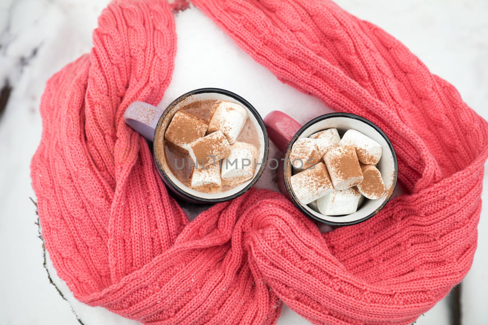 Hot chocolate with marshmallow in pink and violet two cups wrapped in a cozy winter pink scarf on the snow-covered table in the garden. Coloring and processing photo, small depth of field