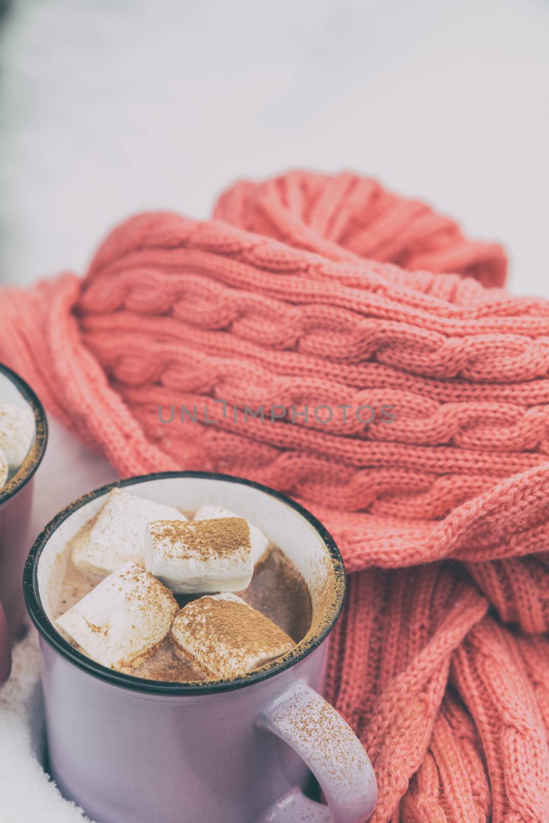 Hot chocolate with marshmallow in violet cup wrapped in a cozy winter pink scarf on the snow-covered table in the garden. Coloring and processing photo, selective focus, small depth of field