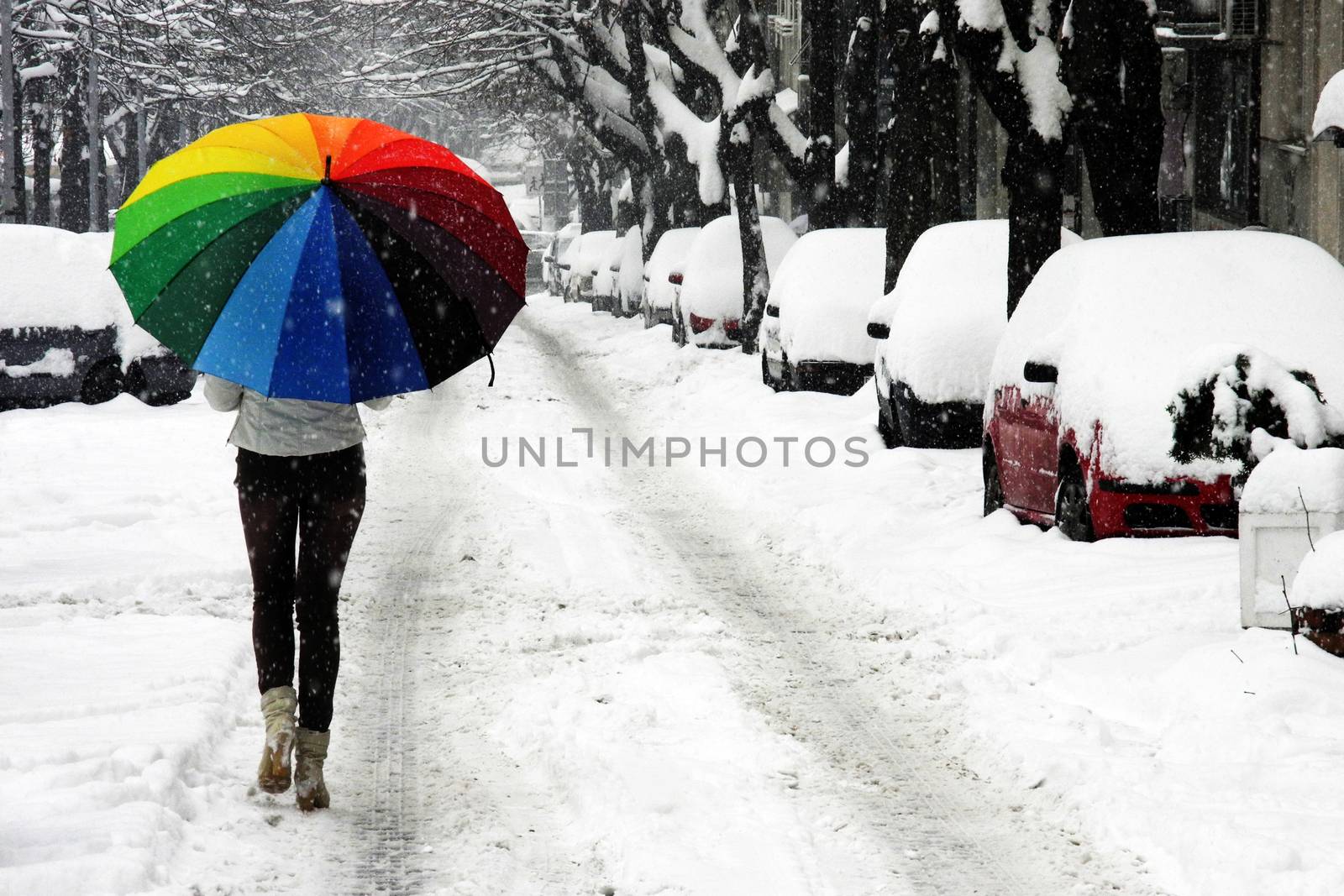 Young woman with rainbow umbrella walking on the street covered with snow