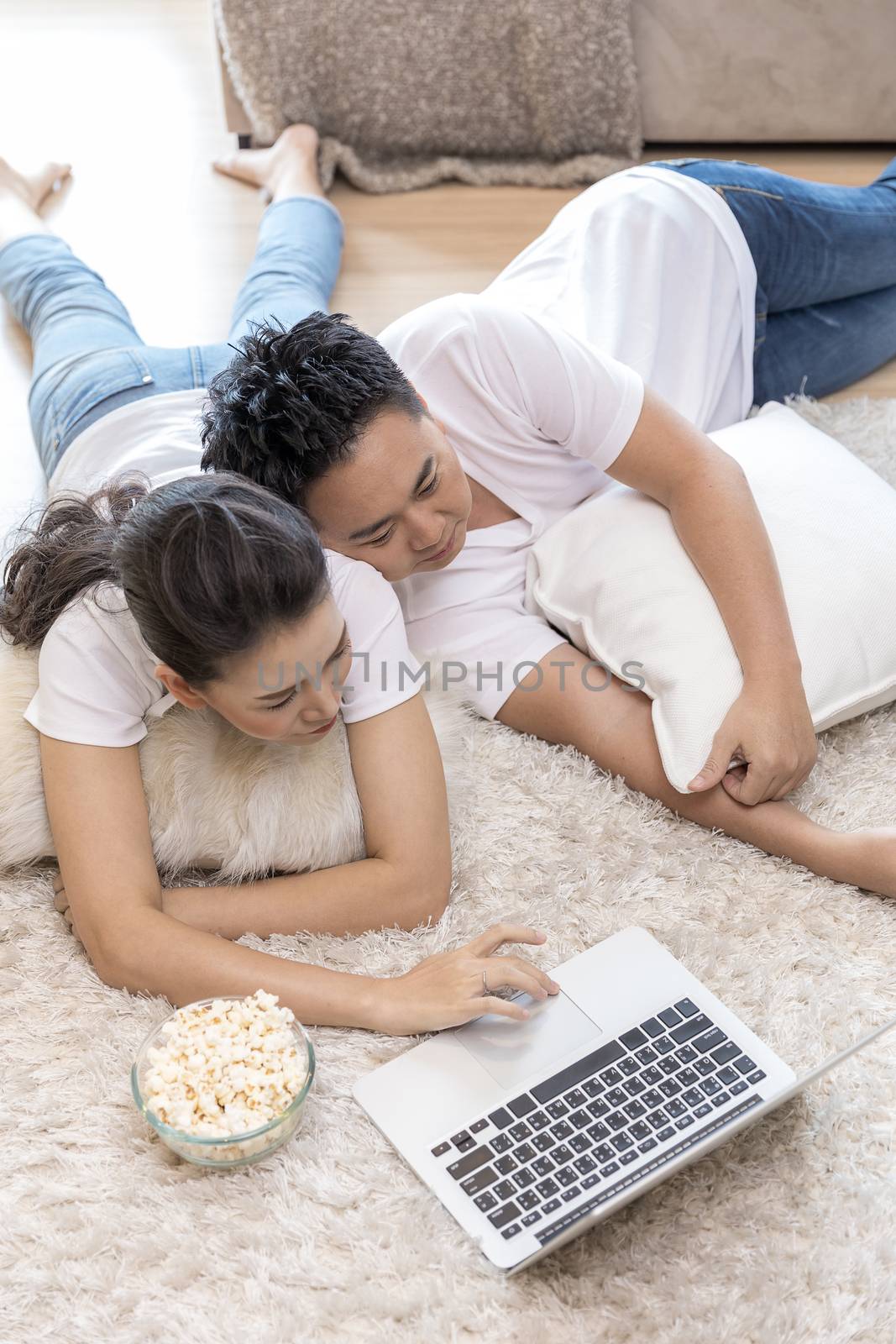 Young Asian Couples lying down and using laptop together in living room of contemporary house for modern lifestyle concept