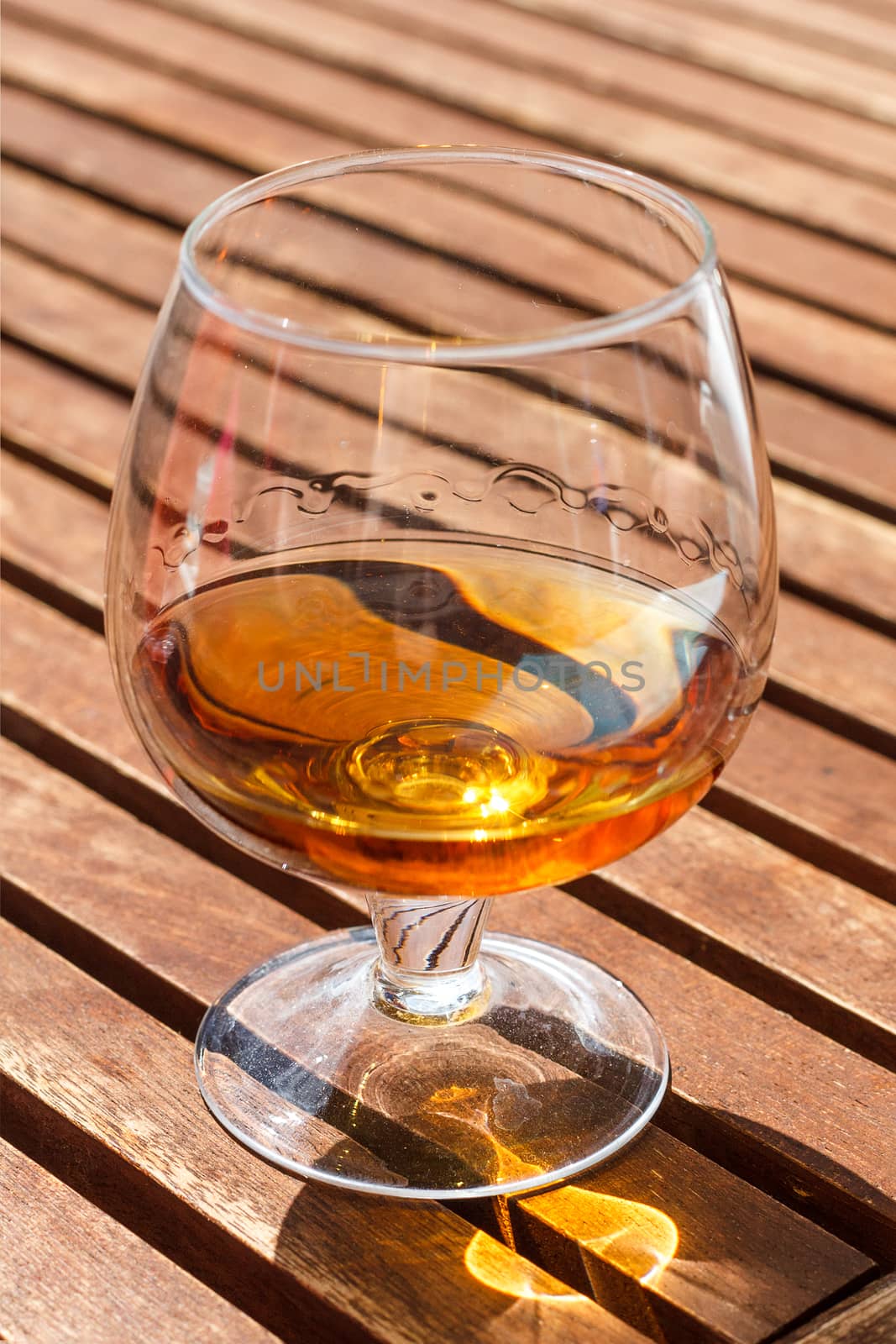 Glass of cognac standing on a wooden table in sunlight.
