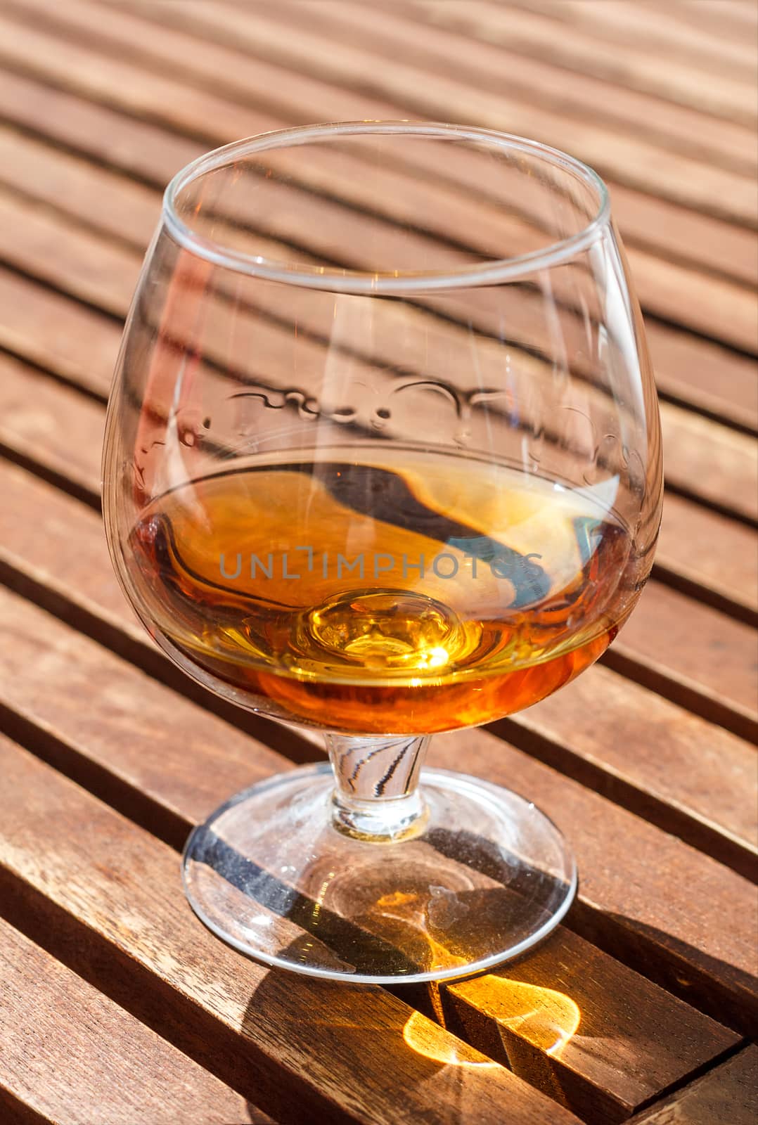 Glass of cognac standing on a wooden table in sunlight.
