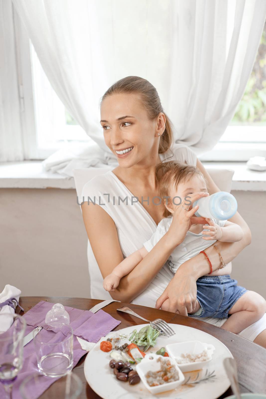 A happy smiling mother feeding her baby with milk bottle in a restaurant.