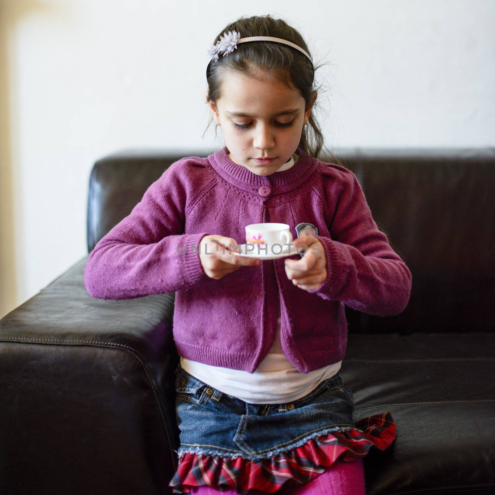 little girl plays, drinking tea on the sofa at home, lit by the window