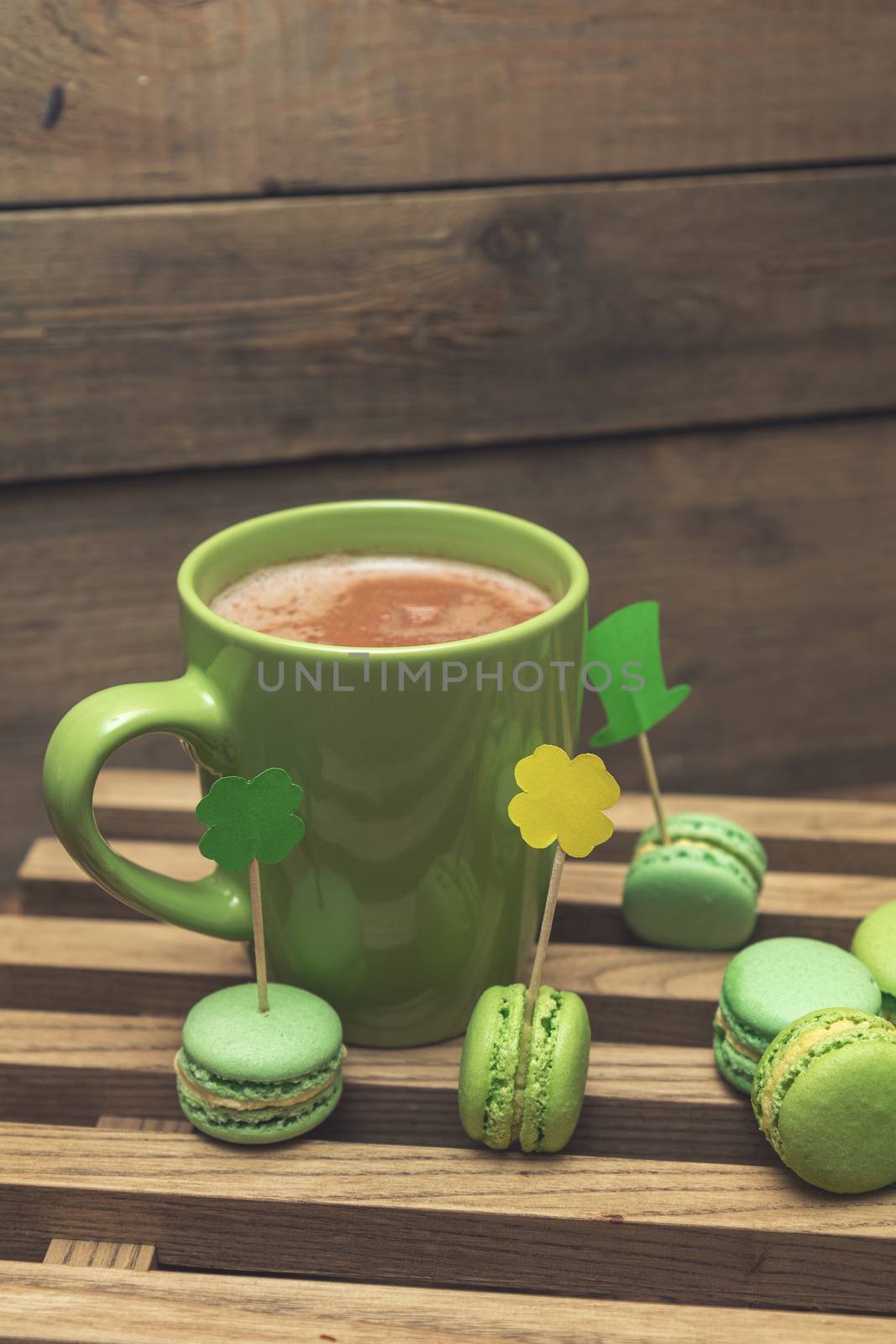 Hot cocoa in green cup and green macaroon cookies scattered on the wooden surface with St. Patrick’s Day attributes. Tonted photo. Shallow depth of field. Copy space