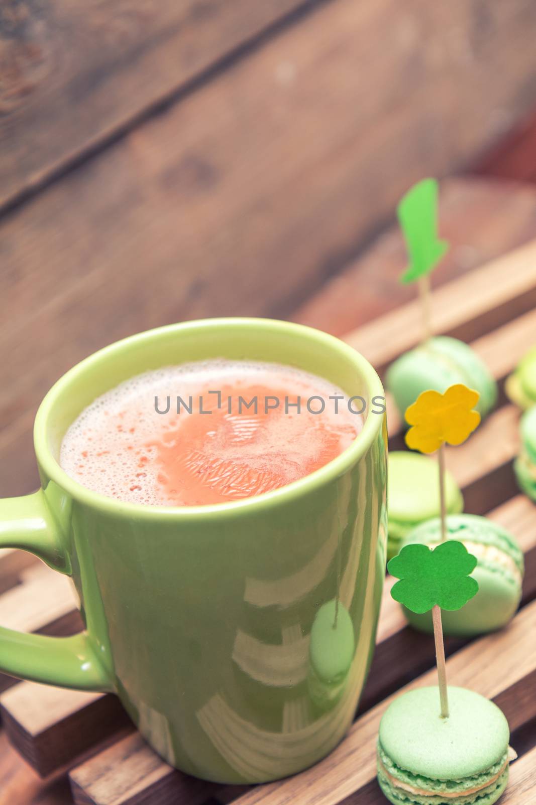 Hot cocoa in green cup and green macaroon cookies scattered on the wooden surface with St. Patrick’s Day attributes. Tonted photo. Shallow depth of field. Copy space