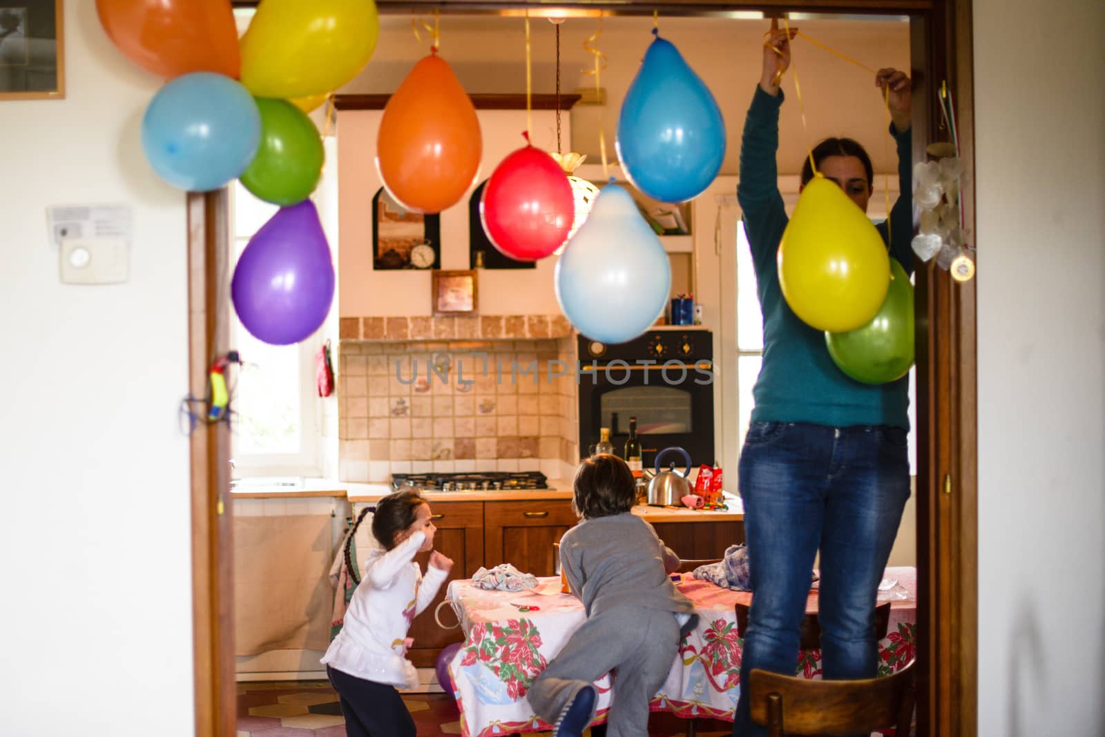 mother with children hang colorful balloons for birthday party in her home, mother standing on the chair hangs balloons at the door