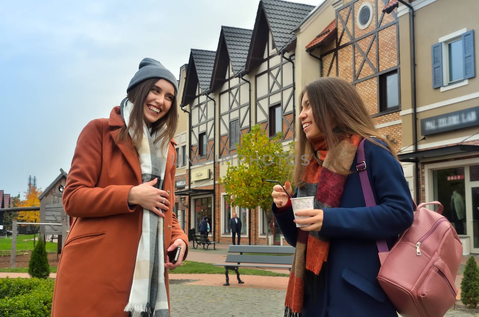 The two girlfriends laugh standing in the street near the shops, with coffee. Dressed in a coat, with backpacks. The cadre is from below. Horizontal photo.