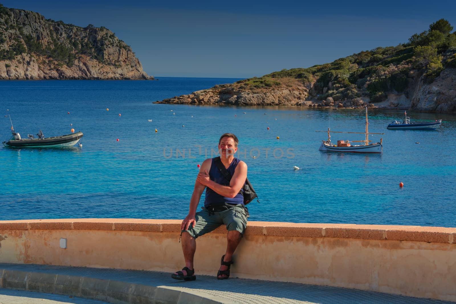 Man sitting on a wall overlooking the side of the sea and relaxing.
Concept of freedom. In the background a rock with a rusty ship anchor. Mallorca, Spain.