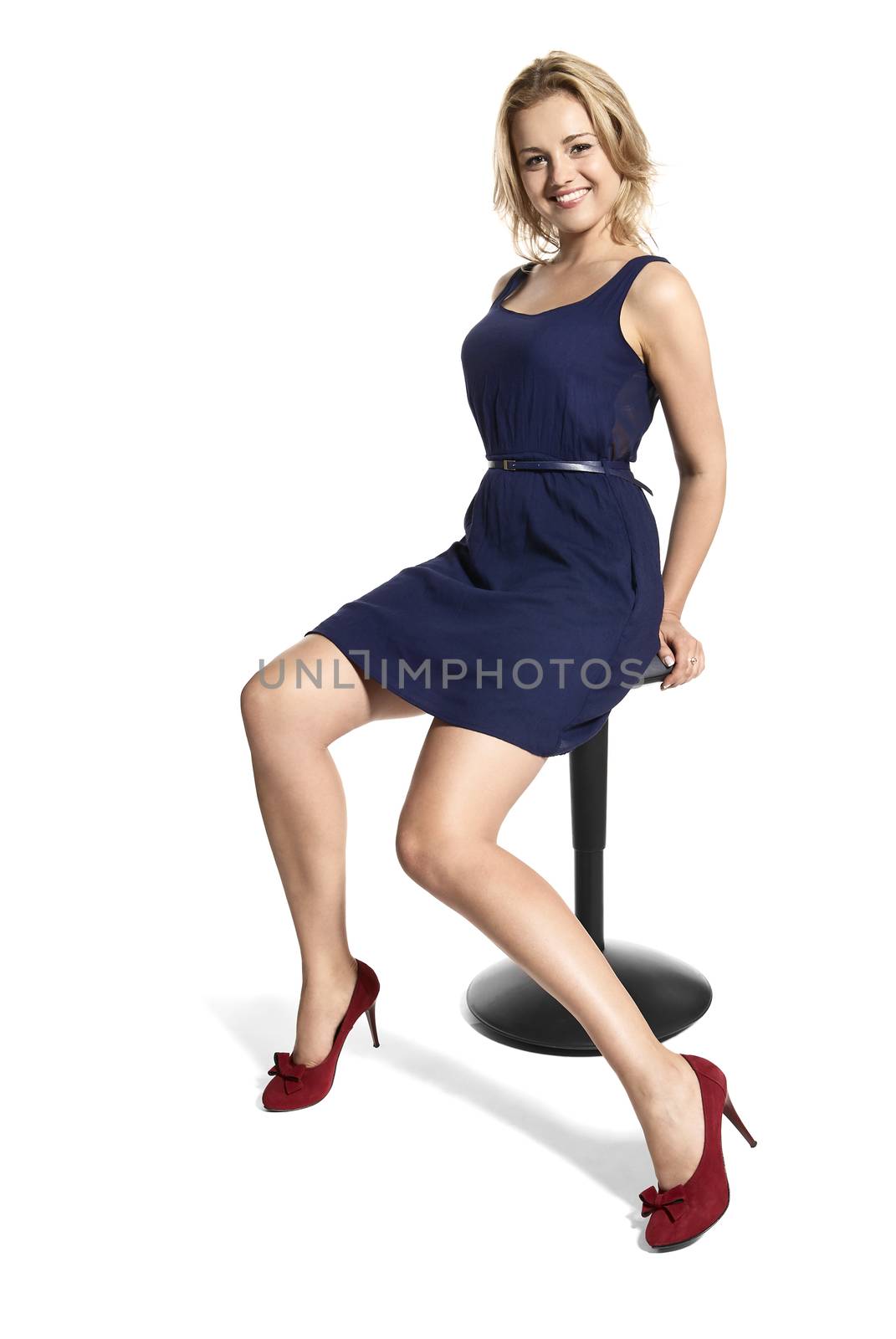 Portrait of sexy young blonde woman in blue dress and red high heels sitting on office stool and smiling to the camera.