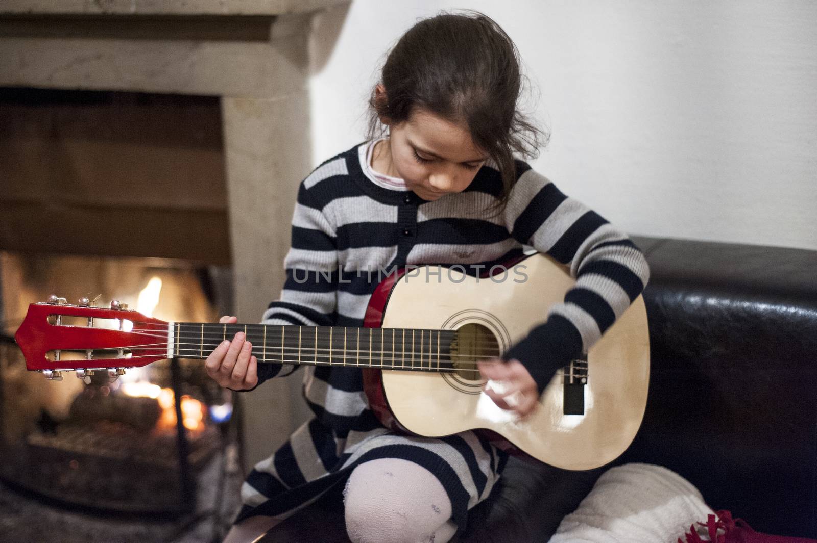 portrait of a 6 year old girl playing guitar at home in front of lit fireplace