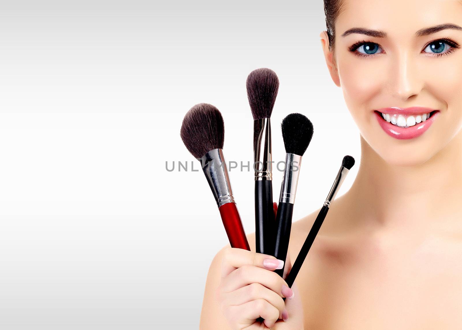 Beauty portrait of lovely beautiful happily smiling woman holding a bunch of make-up brushes against a grey background with copyspace