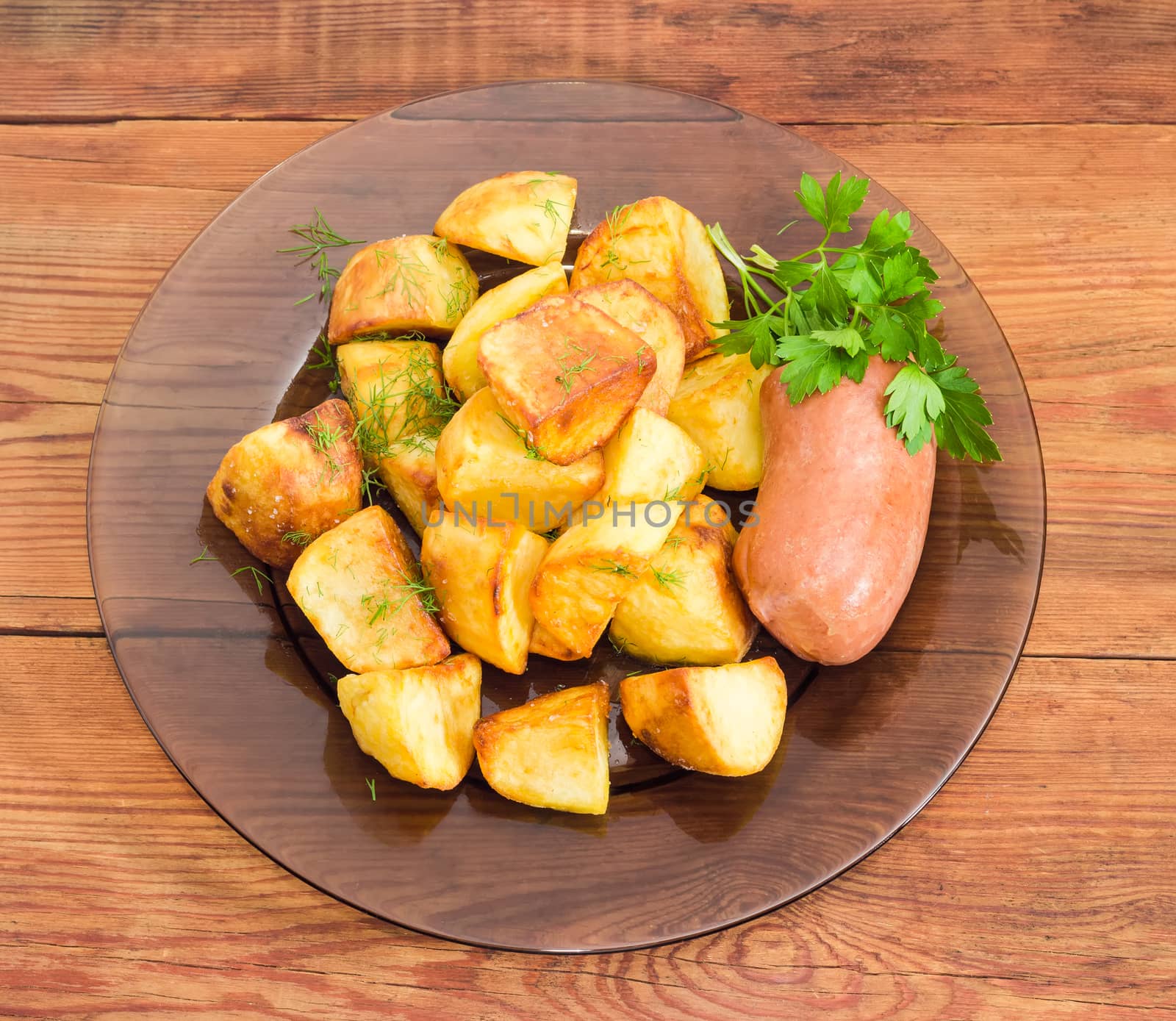 Top view of the dark glass dish with fried potatoes sprinkled by chopped dill, fried wieners and twig of parsley closeup on a surface of old wooden planks
