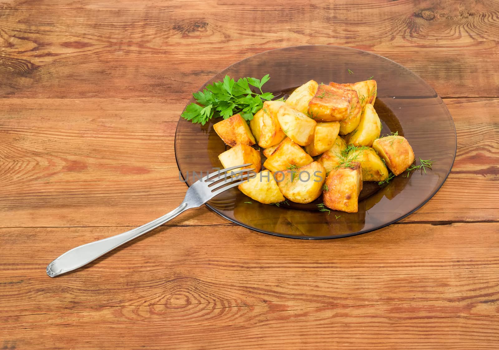 Serving of the country style fried potatoes sprinkled by chopped dill and twig of parsley on dark glass dish and fork on a surface of old wooden planks
