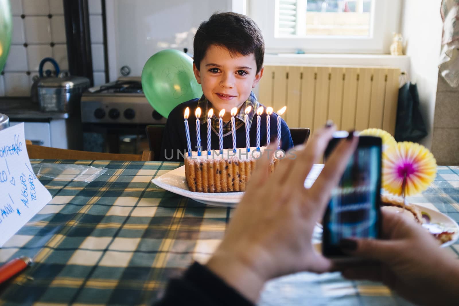 baby on the day of his ninth birthday blowing the candles on the cake, in the kitchen of his home, his mother films him with a smartphone