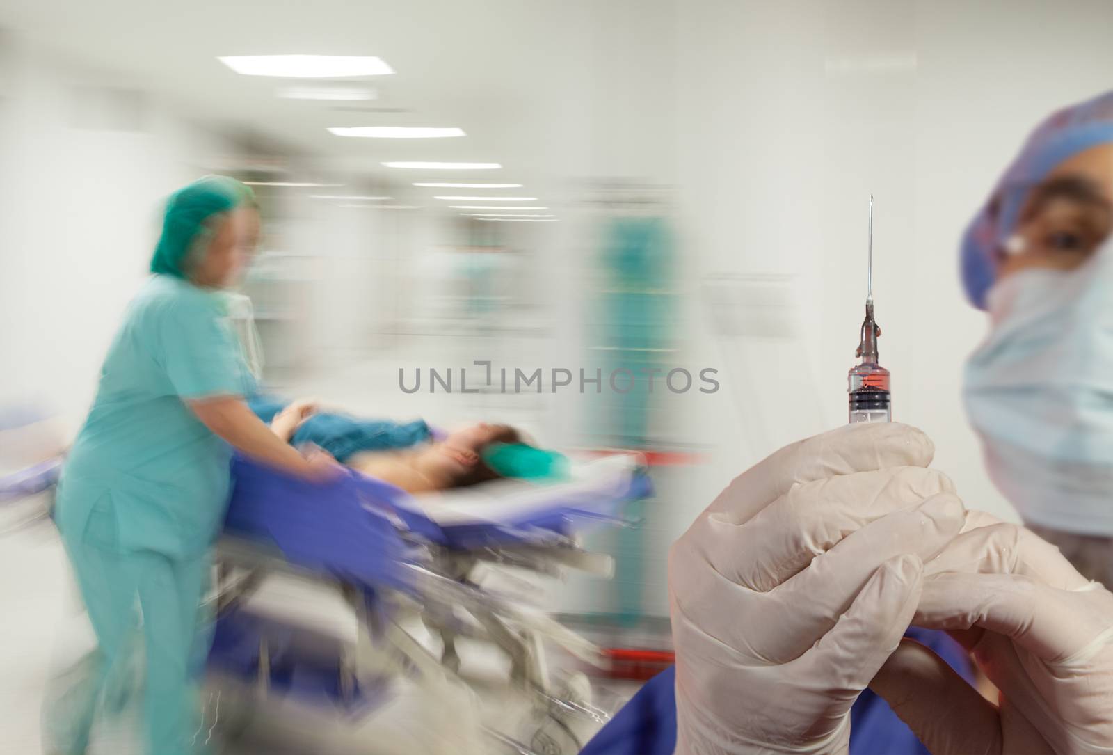 A doctor with syringe and gloves over motion blurred background of nurse in hurry pushing litter down a sterile light hospital corridor.