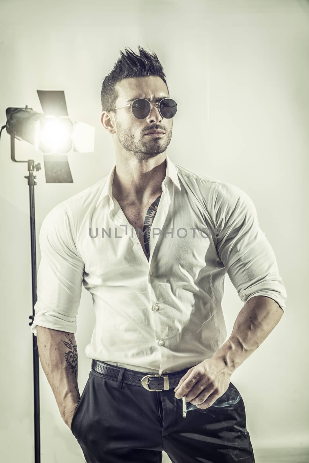 Young macho in formal shirt and sunglasses holding cigarette posing confidently in studio.