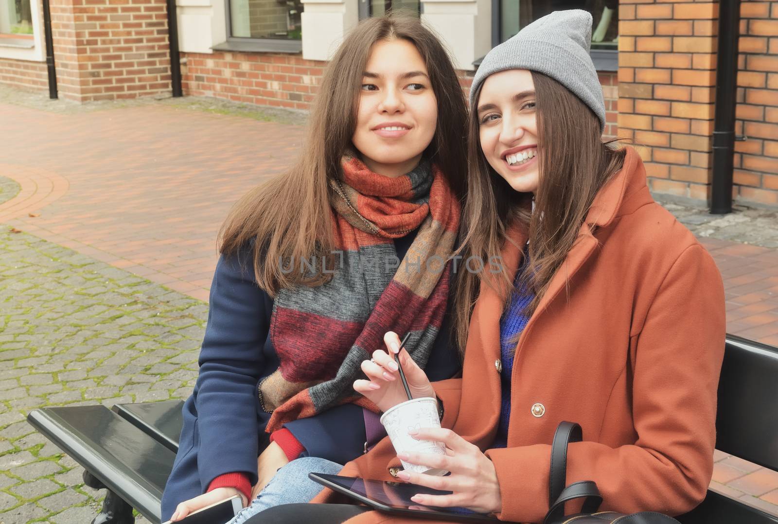 Two girls sit on the bench outside and smile, look at the photographer. Dressed in a coat, hold the phone and tablet in their hands. Asian and European
