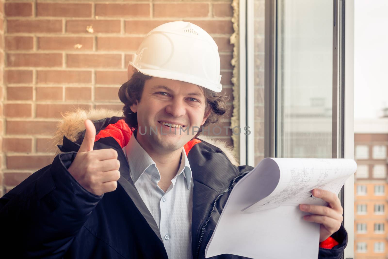 Handyman giving thumbs-up. Engineer with blueprint on brick background. Happy builder with pleased smile. Soft focus, toned